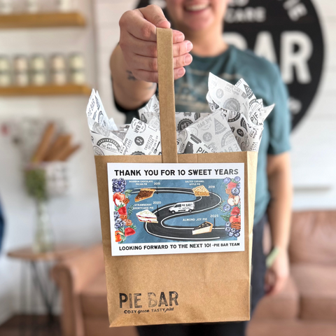 Person holding a brown paper bag with 'Pie Bar' branding and celebratory text.