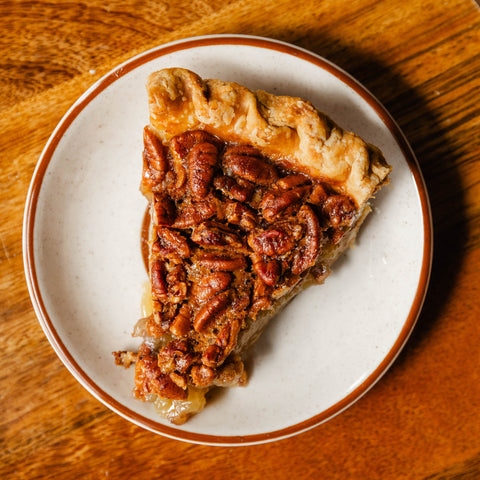 Slice of pecan pie on a white plate with a brown rim, placed on a wooden surface.