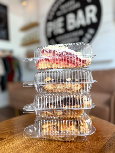 Stack of pie slices in clear plastic containers on a wooden table with 'Pie Bar' sign in the background.
