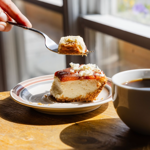 Slice of cheesecake on a plate with a cup of coffee, held by a hand with a fork.