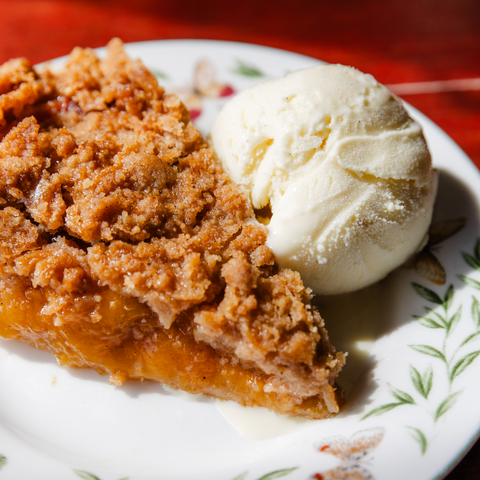 Slice of peach pie with crumble topping and a scoop of vanilla ice cream on a decorative plate.
